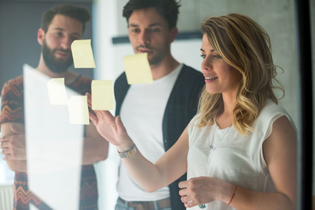 Zwei Männer und eine Frau stehen vor einer Glasscheibe, auf der drei gelbe Post-Its kleben